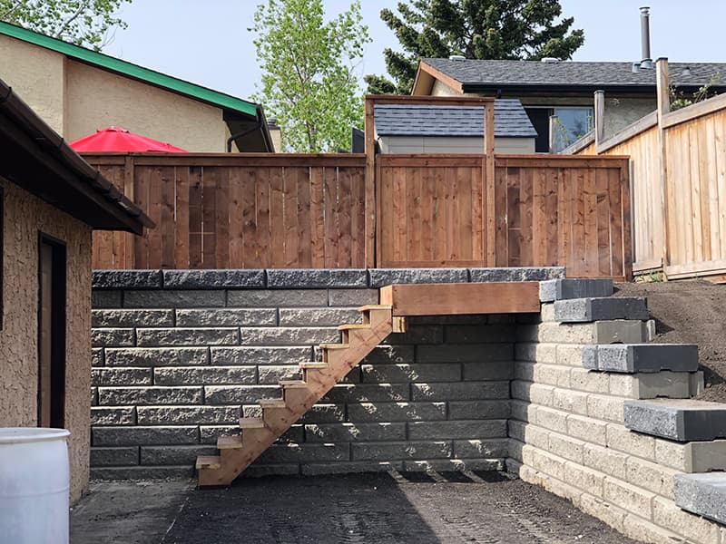 KBS Contracting & Bobcat Services image content 1 Backyard with a stone retaining wall, wooden fence, and stairs, showcasing the work of KBS Contracting & Bobcat Services, expert retaining wall contractors in Calgary.
